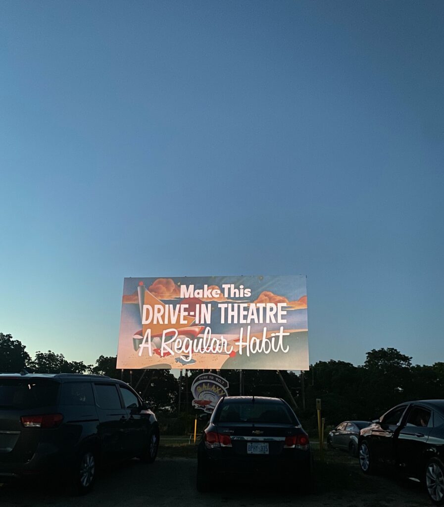 An evening at a drive-in theater with a sign recommending the viewers make it a habit